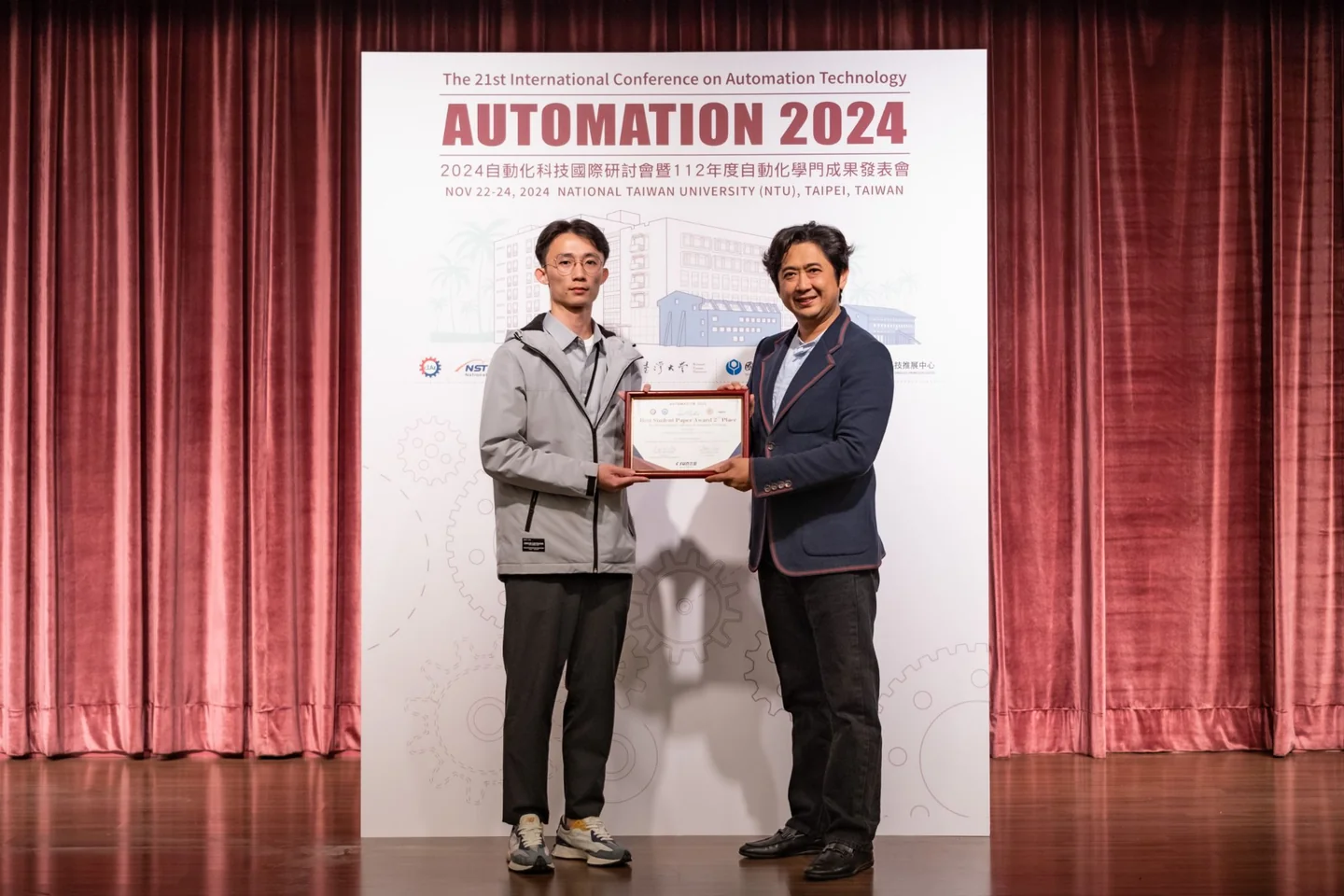 頒獎人與得獎人在 AUTOMATION 2024 主視覺看板前共同持獎狀合影。