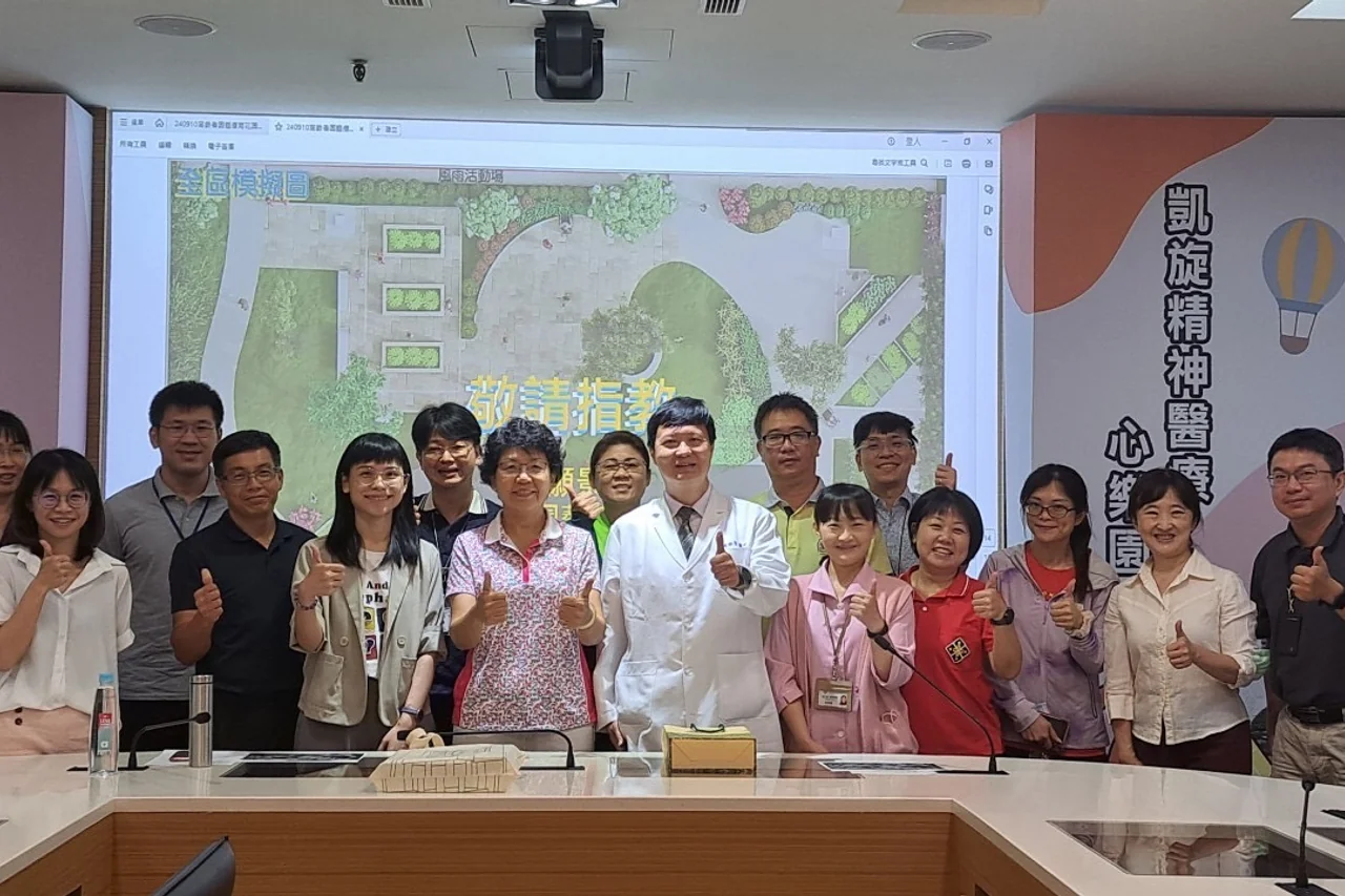 會議室內十餘人站成一排比出讚的手勢，背後投影幕顯示園區全區模擬圖。