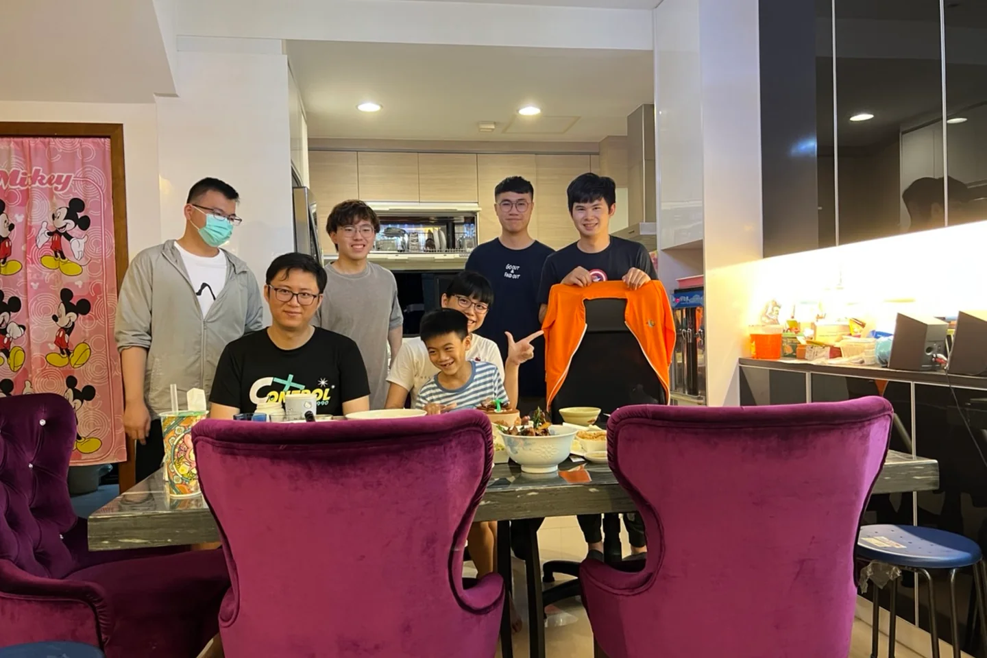 室內餐桌旁七人合影，桌上擺著餐點，其中一人展示一件橘色外套。