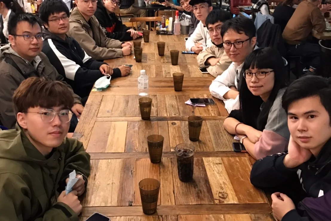餐廳長桌兩側坐著十餘位成員，全員轉頭看向鏡頭，桌上擺著水杯與飲料。
