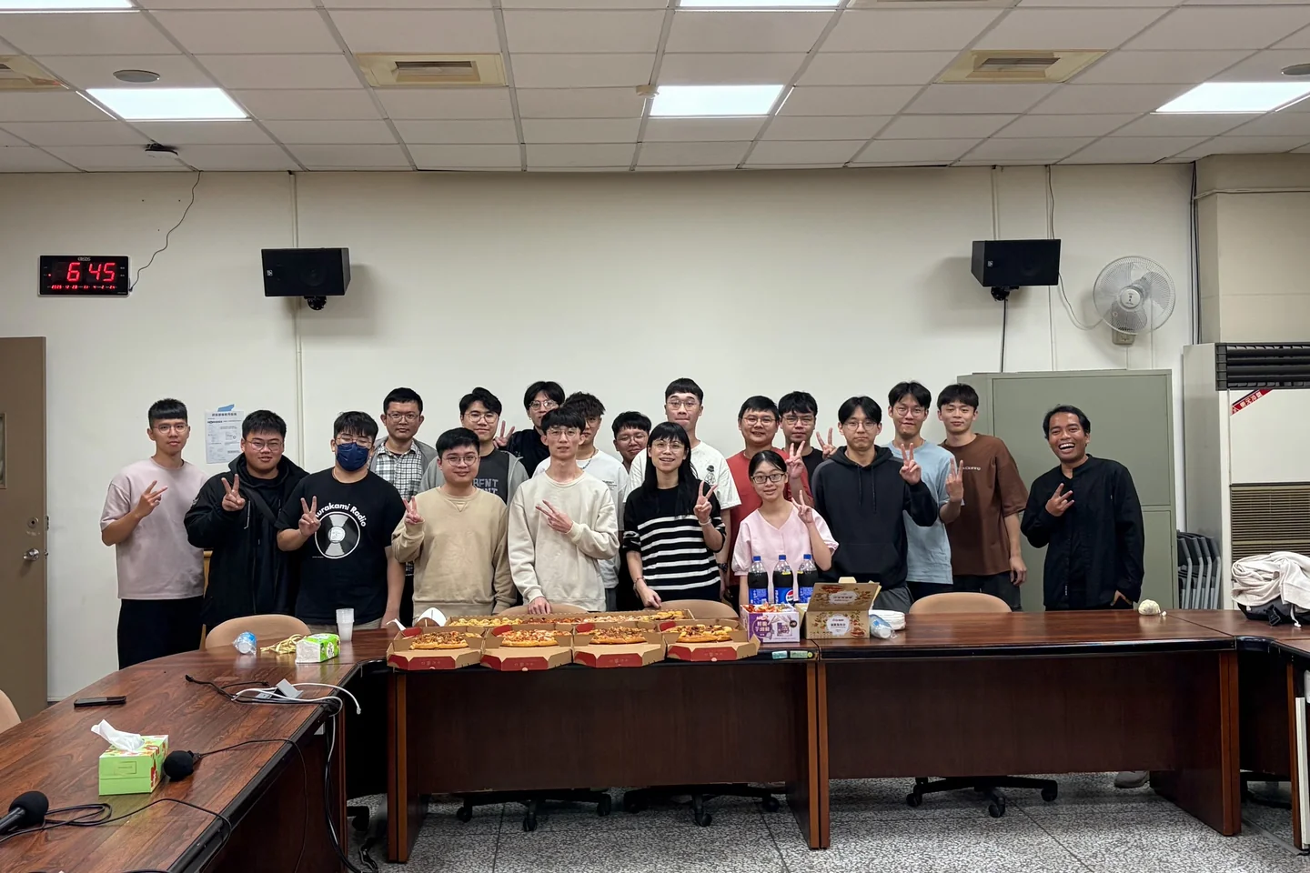 研討室內約二十位成員站在長桌後方合影，桌上擺著四盒披薩與飲料。