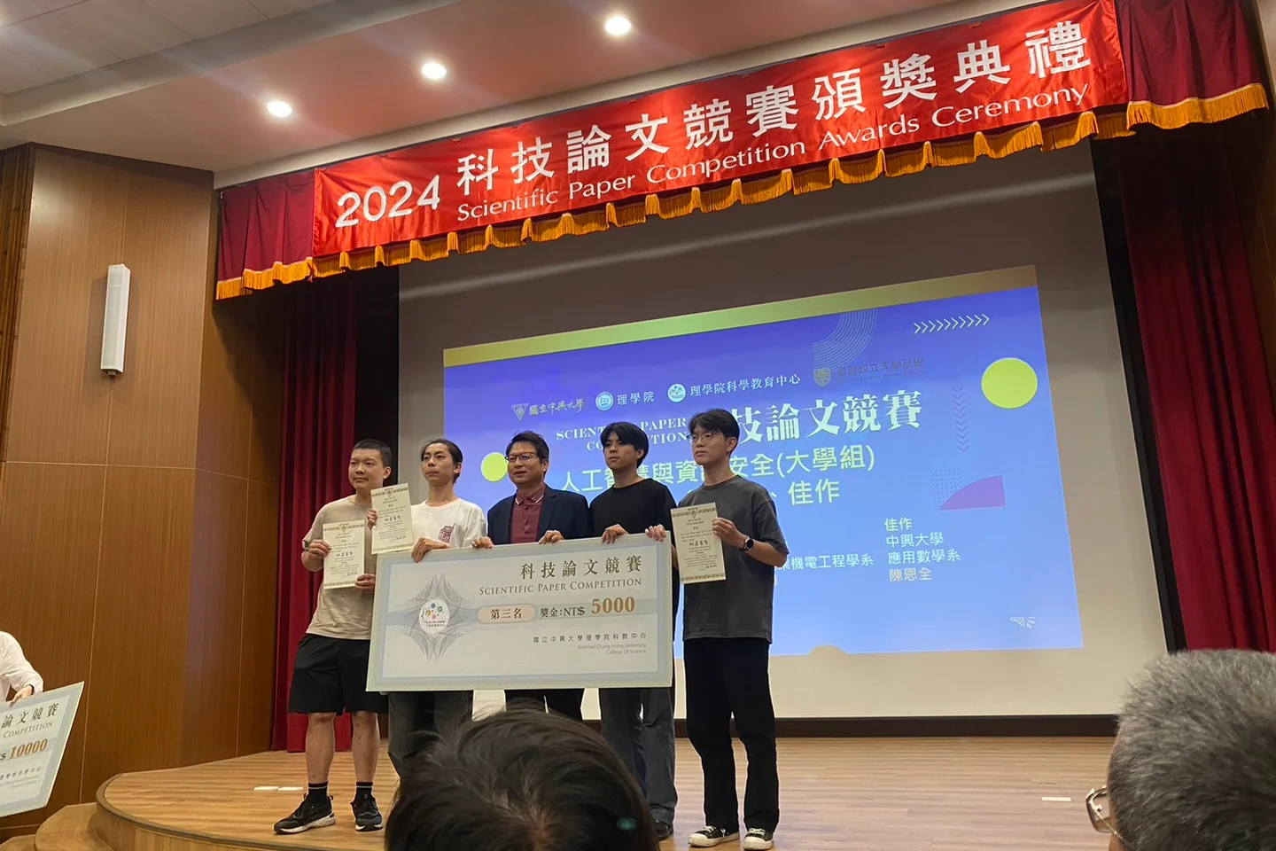 頒獎典禮舞台上，得獎人手持獎狀與第三名獎金看板合影，背後紅色橫幅寫著科技論文競賽頒獎典禮。