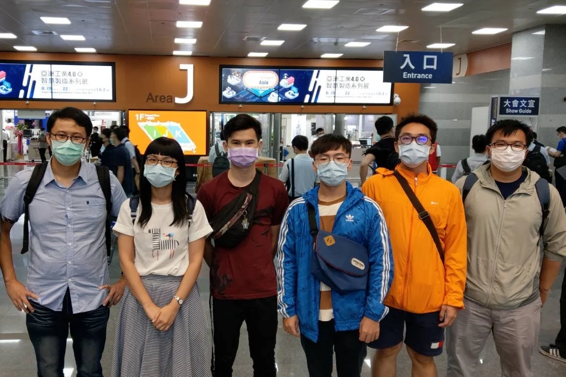 展館入口前六人合影，全員戴口罩，背後電子看板顯示展覽名稱與 Area J 入口指示。